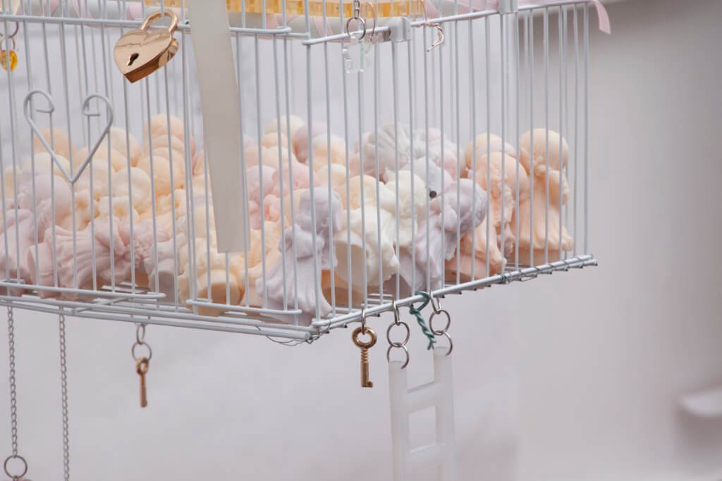 Das Kunstwerk "White Suburban Home" von Emma Pryde. Man sieht einen Vogelkäfig voller pastellfarbener Engelsfiguren. Am Käfig sind ein Schloss, Schlüssel und eine Leiter befestigt.