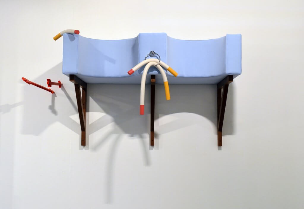 Das Kunstwerk "Tongue-tied" von Liam Fallon. Man sieht eine hellblaue Wandskulptur, die mit Zigaretten versehen ist, links daneben zwei rote Pfeile.