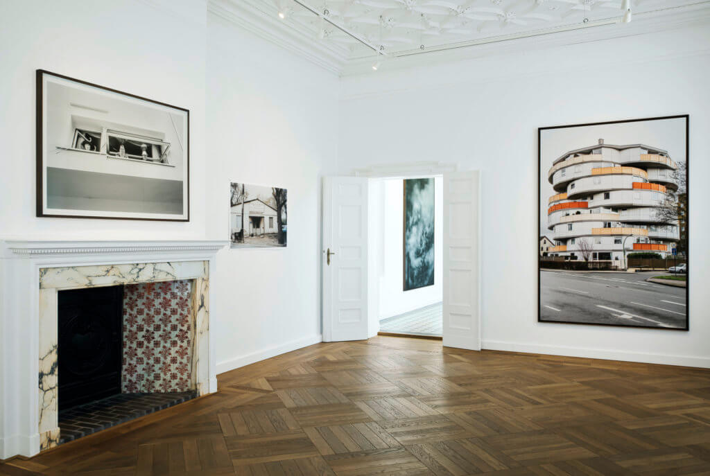 Ausstellungsansicht aus der Villa Schöningen. Ein Raum mit Parkettboden und Kamin, an den Wänden Fotografien.