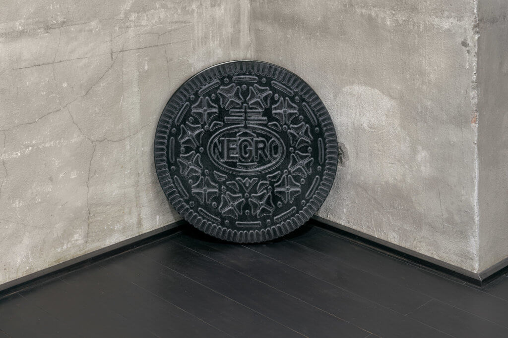 Eine Arbeit des Künstlers Michael Sayles. Zu sehen ist ein kreisrundes Bild, das an einen Oreo-Keks erinnert. Statt des Markennamens ist "NEGRO" zu lesen.