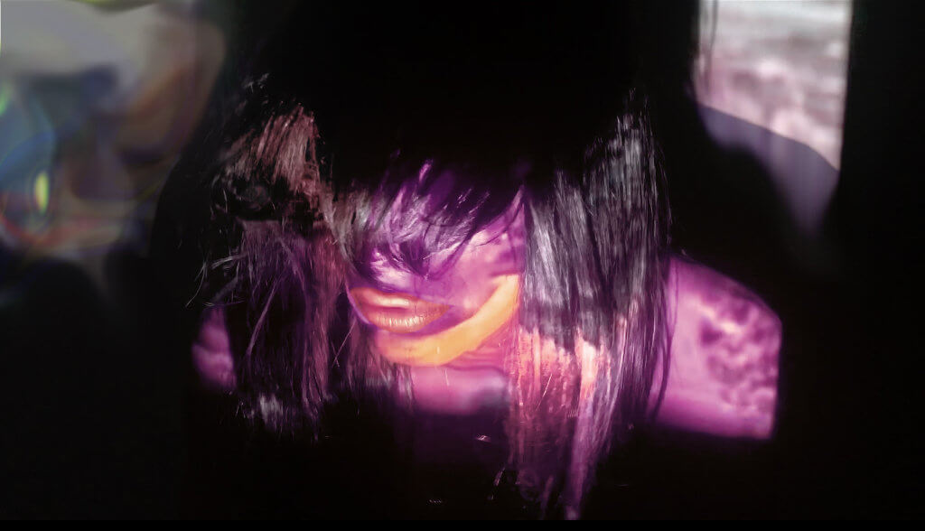 Das Bild zeigt den Künstler MJ Harper während einer Performance. Der Ausschnitt zeigt lediglich den Kopf und die Schultern des Künstlers, beide sind mit violetter Farbe beschienen, zudem trägt der Künstler eine Langhaarperücke. Das Bild gibt keine spezifische Auskunft über die Performance.