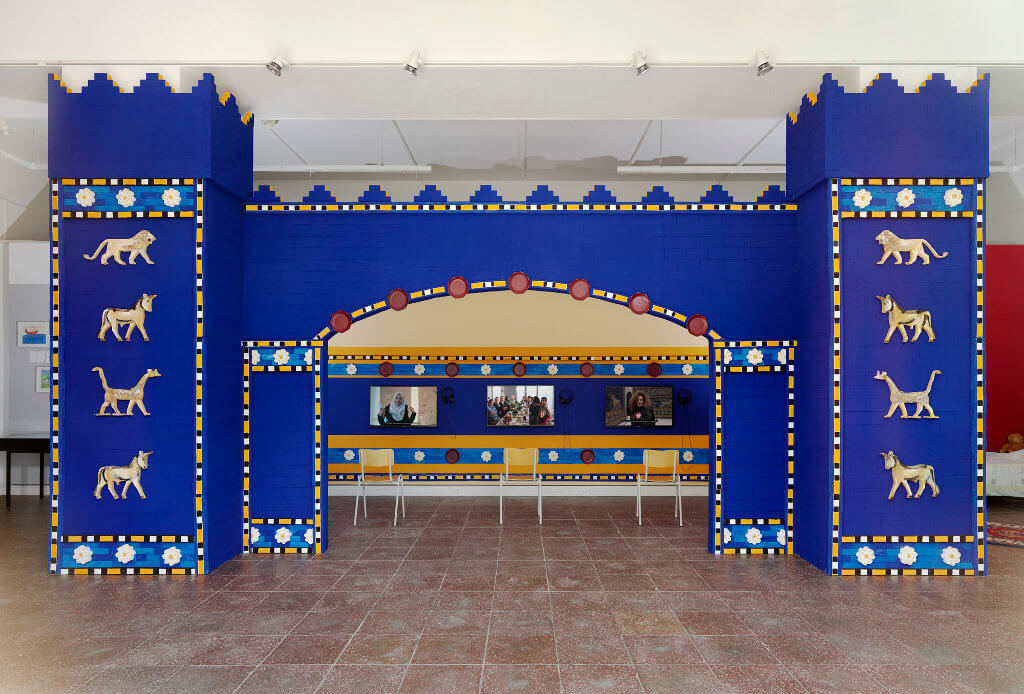 Das Bild zeigt eine Ausstellung-Ansicht der Ausstellung "Fasahat". Zu sehen ist eine Nachbildung des Ishtar-Tor mit drei Bildschirmen im Hintergrund. Das Tor hat eine kräftige blaue Farbe.