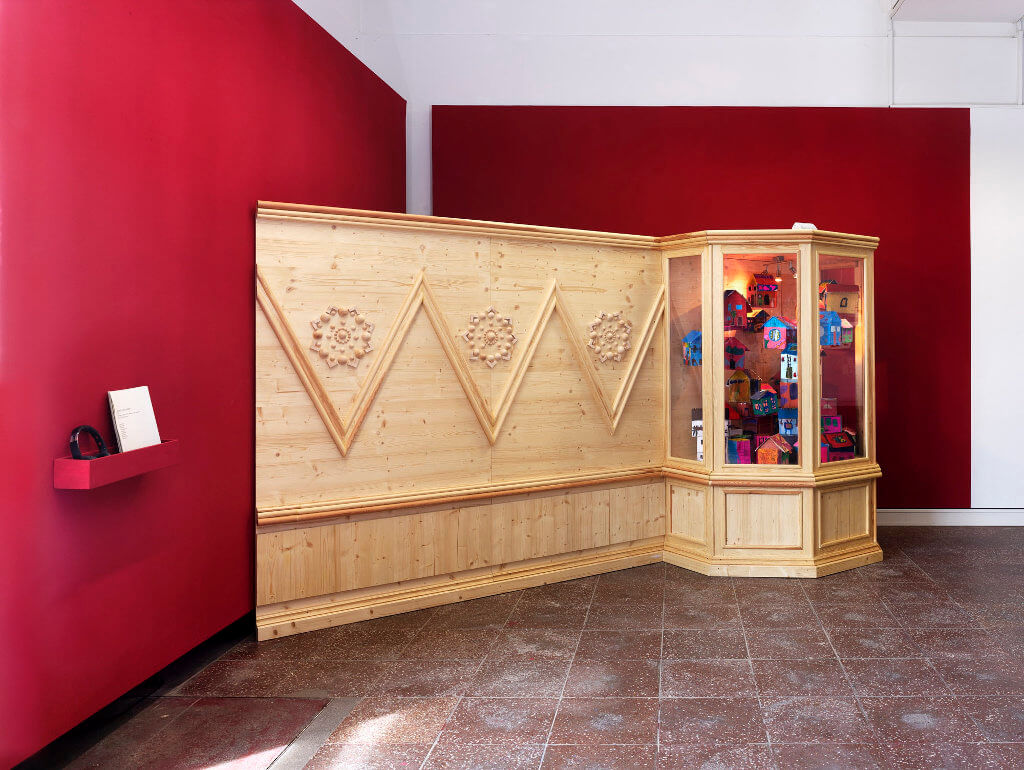 Das Bild zeigt eine Ausstellung-Ansicht der Ausstellung "Fasahat". Zu sehen ist eine verzierte Holzwand mit einer Vitrine an einem Ende. Die Vertäfelung steht vor einer roten Wand, es handelt sich um eine Installation.