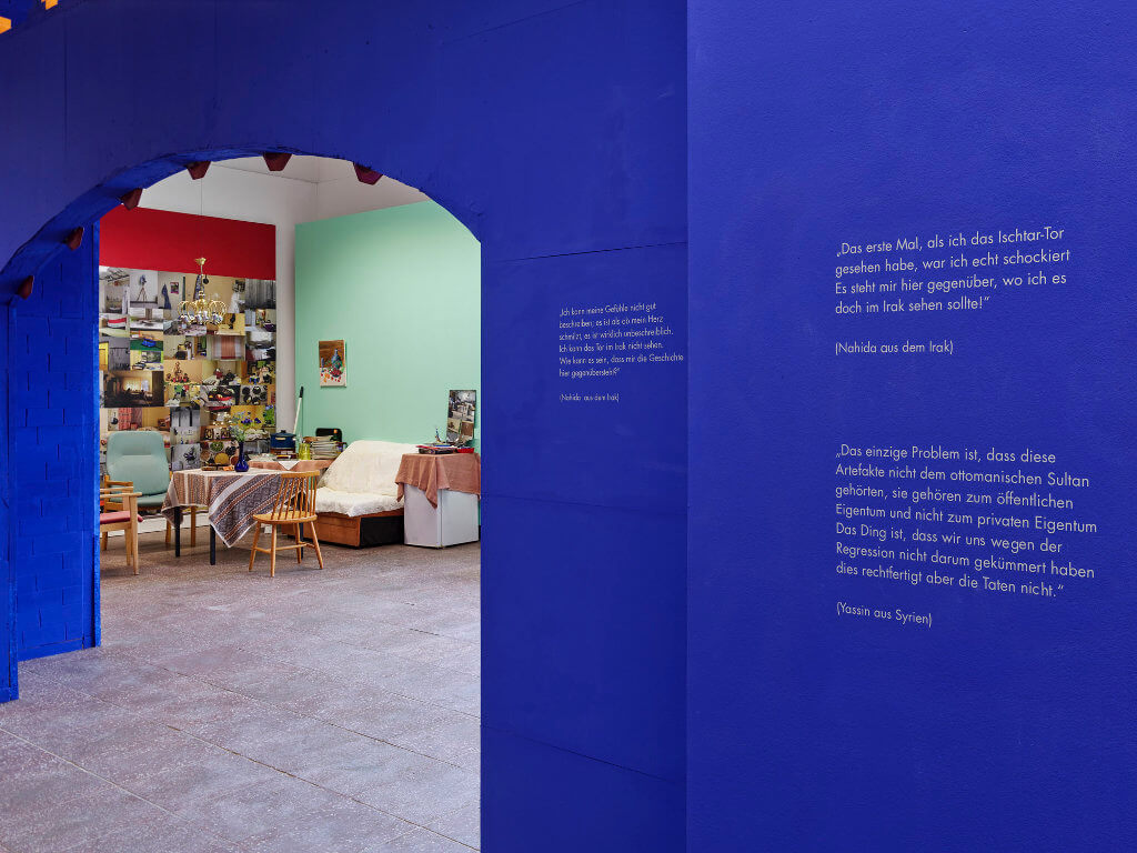 Das Bild zeigt eine Ausstellung-Ansicht der Ausstellung "Fasahat". Zu sehen ist die Rückwand des nachgebauten Ishtar-Tors in blauer Farbe mit Zitaten darauf gedruckt.