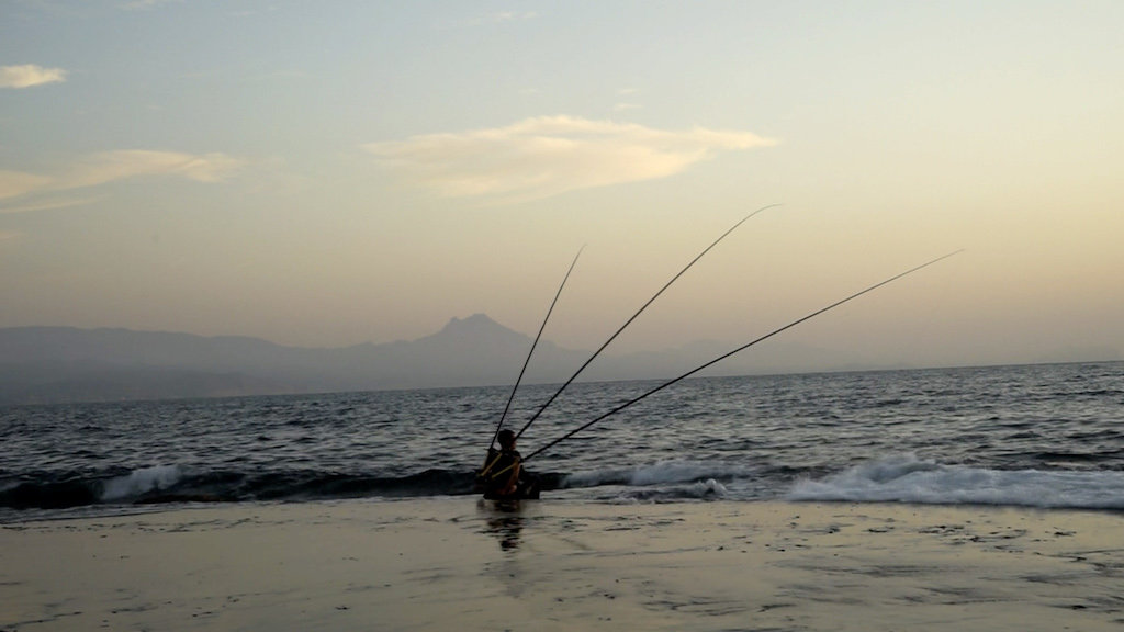 Filmstill aus ‚Transmission Waves’ von Judith Egger zeigt die Künstlerin mit ihrer Konstruktion aus Angelruten auf dem Rücken im Meer
