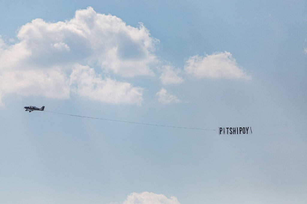 Das Bild zeigt ein Flugzeug vor einem blauen Himmel, welches ein Banner mit der Aufschrift "Pitshipoy" nach sich zieht.