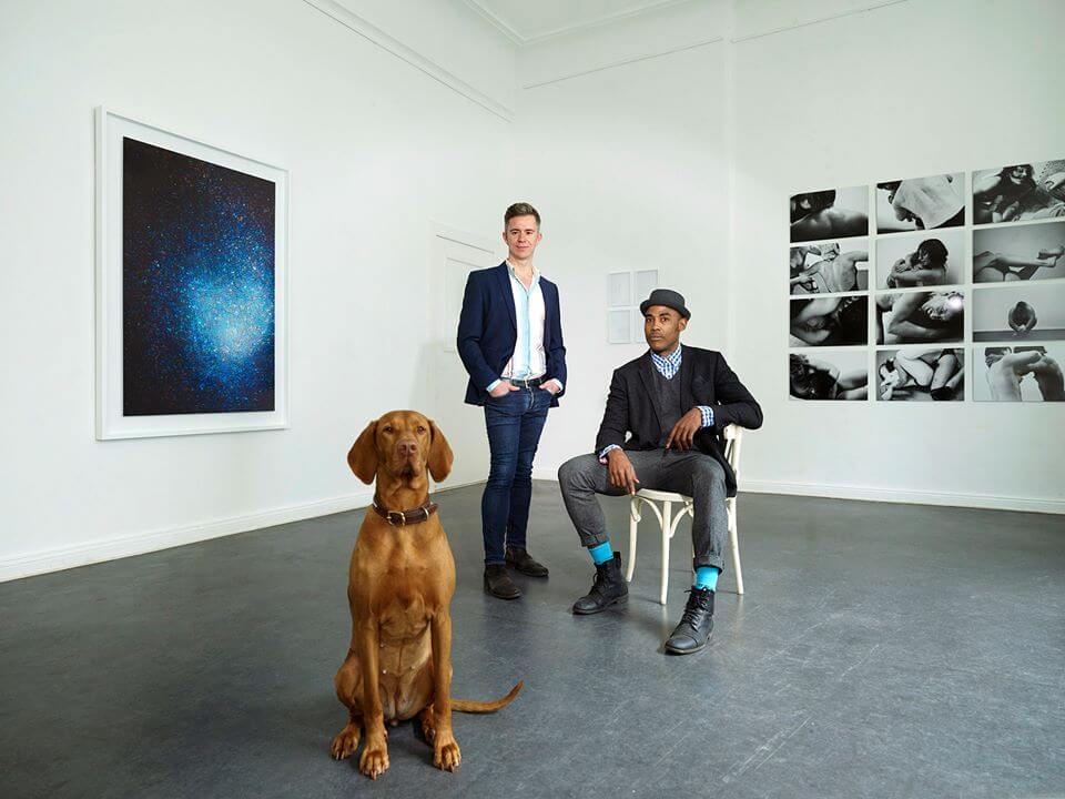 Zwei Männer mit einem Hund in einem weißen Raum.