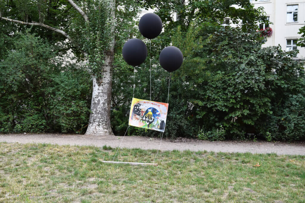Ausstellungsansicht "Ok Park" im Berliner Auerpark. Ein Bild wird von drei schwarzen Luftballons in der Luft gehalten. Eine Arbeit von Anne Duk Hee Jordan: „Lord of the wizard — Last judgement“.