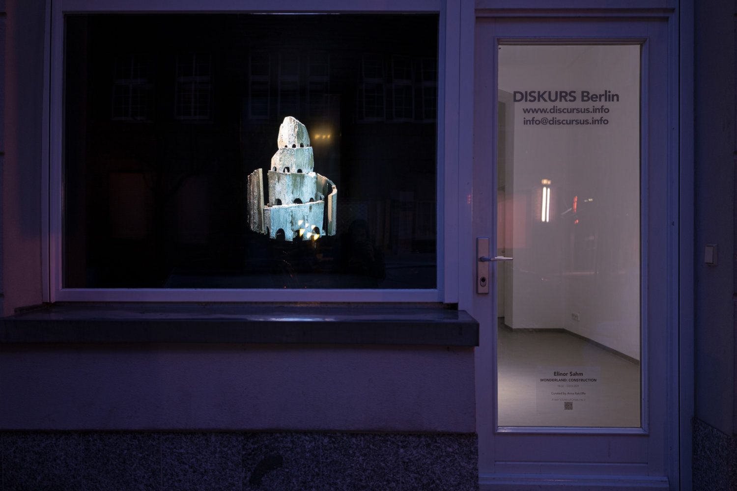 Schaufenster-Ausstellung bei Diskurs Berlin, links ein großes Fenster, wo ein Turm aus Beton zu sehen ist, rechts eine Tür, wo helles Licht erkennbar ist
