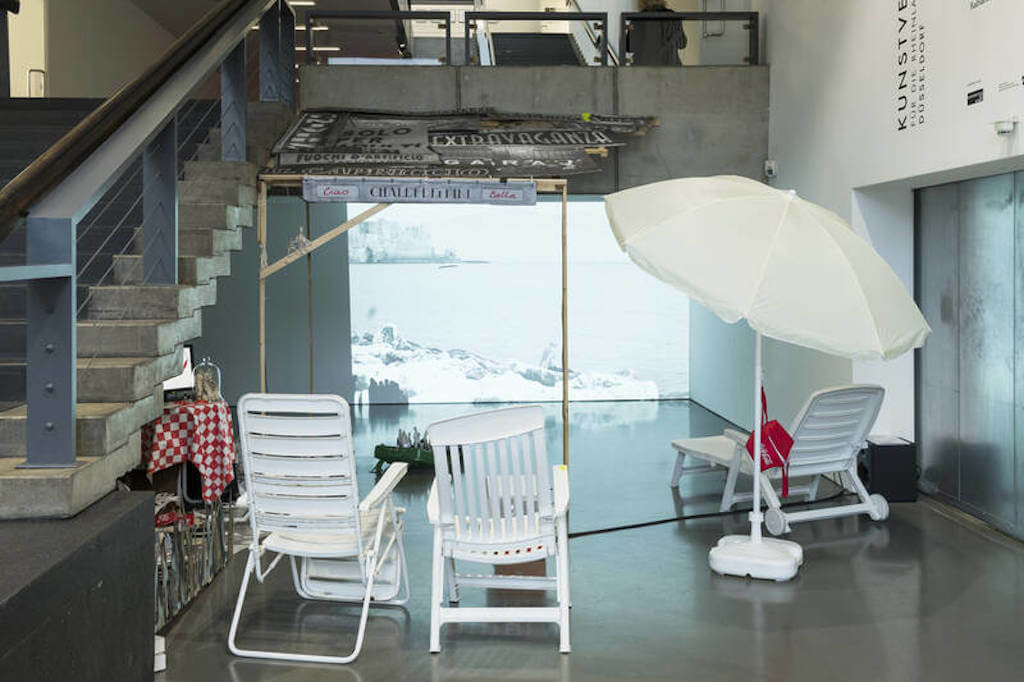 Zu sehen ist eine Installationsansicht der Arbeit " L`estate scorsa" von Serena Ferrario in der Kunsthalle Düsseldorf aus dem Jahr 2019. Man sieht Strandstühle und einen Sonnenschirm vor einer Video-Leinwand.