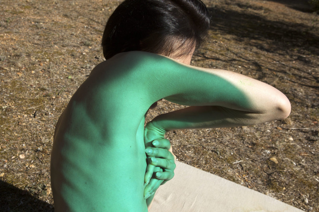 Eine nackte Frau, teilweise mit grüner Farbe bedeckt, verschränkt die Hände vor der Brust, ihr Gesicht ist verdeckt.