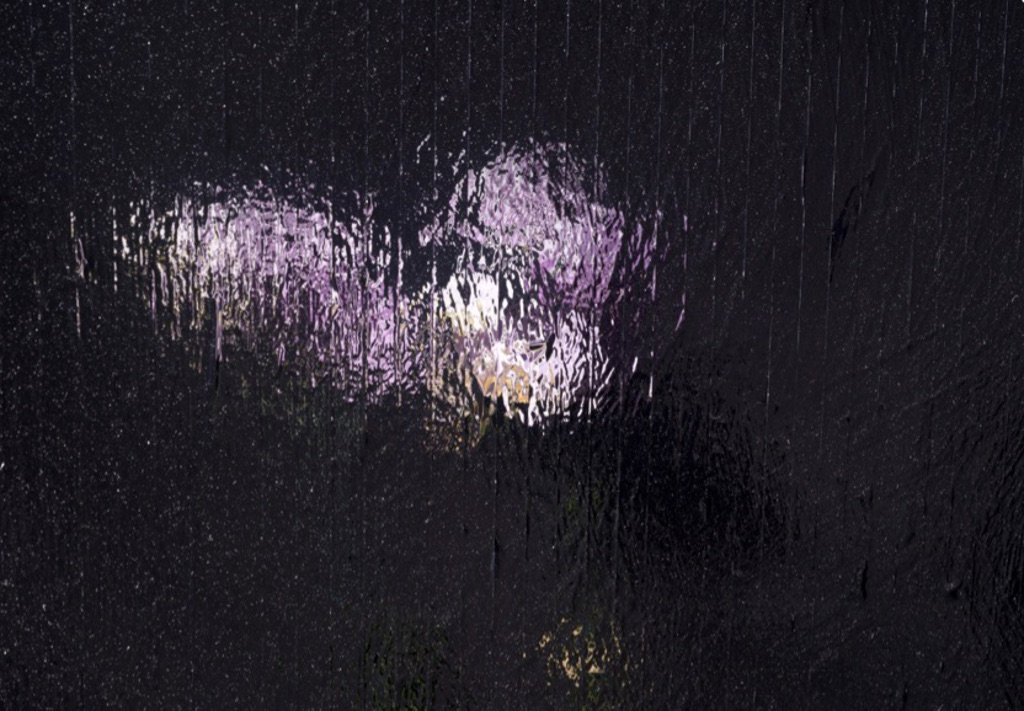Das Bild zeigt eine fliederfarbene Fläche auf schwarzem Untergrund. Der visuelle Eindruck scheint wie eine durch ein Regenfenster aufgenommene Orchidee.
