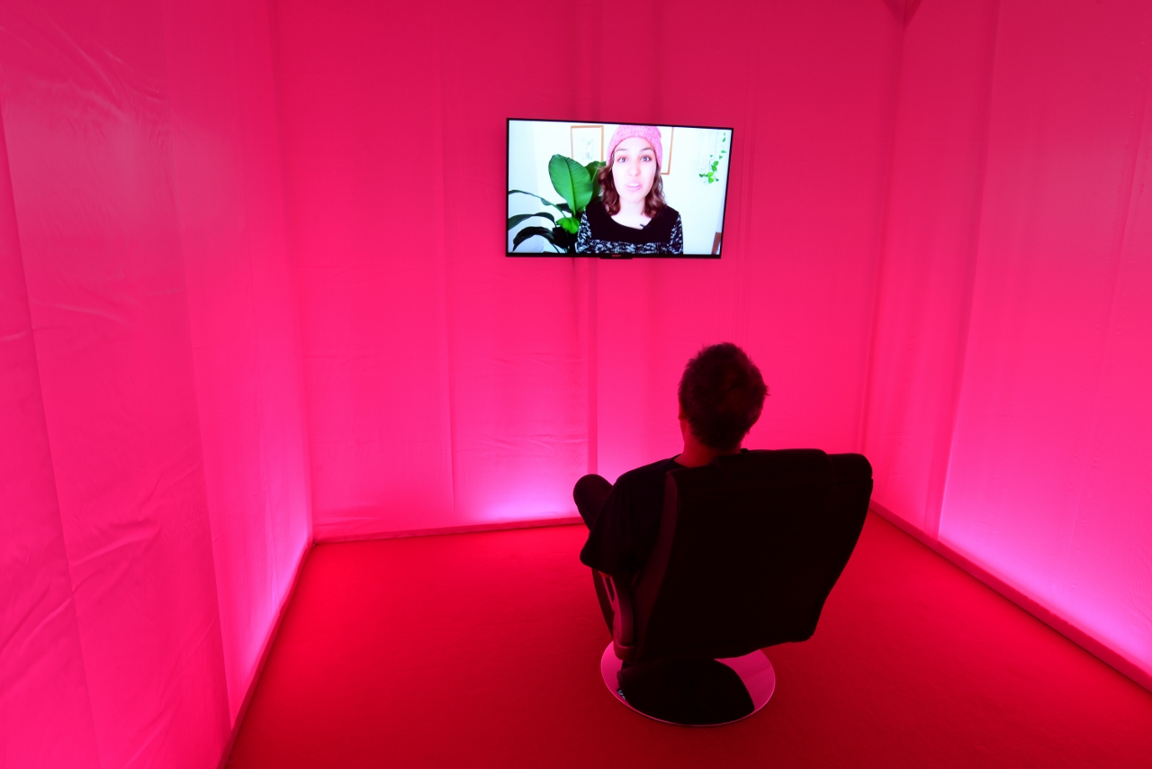 Eine Installation mit Video von BRR mit dem Titel "Self Care Center", zu sehen im Kunstverein Wolfsburg, auf einem Sessel sitzt ein Mann, den Blick auf den Bildschirm gerichtet, wo eine Frau zu sehen ist, die gerade redet, die Raumgestaltung ist leuchtend rot-pink