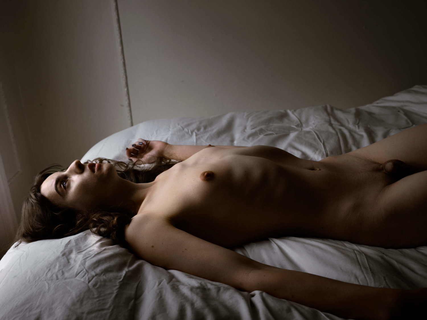 Eine nackte Transfrau liegt mit dem Rücken auf dem Bett, auf einer weißen Bettdecke, die Arme sind ausgestreckt und der Blick nach oben zur Decke gerichtet, auf das Gesicht und den Oberkörper fällt Licht