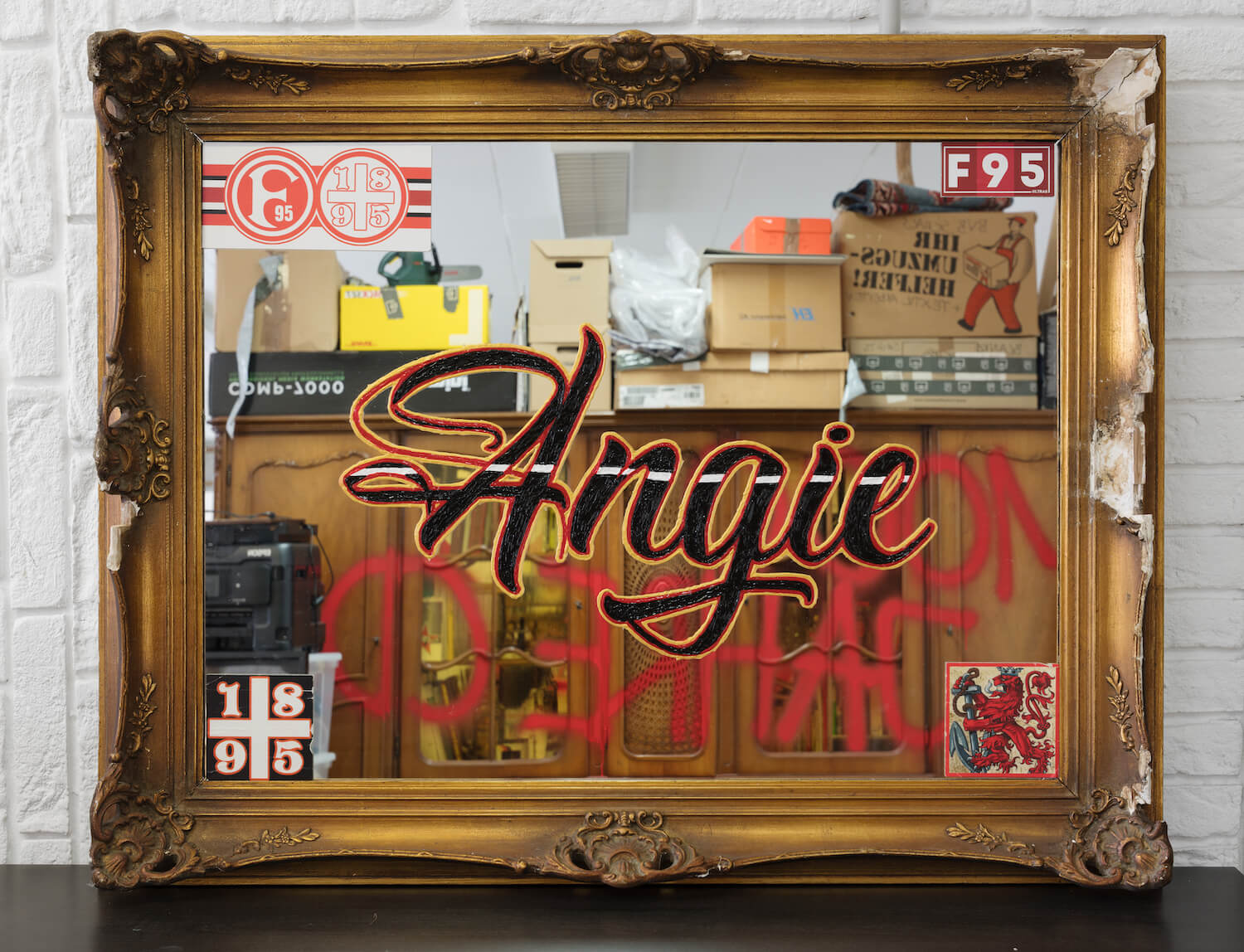 Ein kitschig gerahmter Spiegel mit verschiedenen Augklebern. In der Mitte in Schwarz-rot-gold der Schriftzug "Angie".