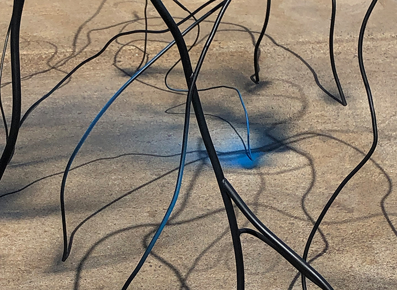 Ein Gebilde aus schwarzem Stahl steht auf Betonboden, auf eine Stelle fällt blaues Licht.