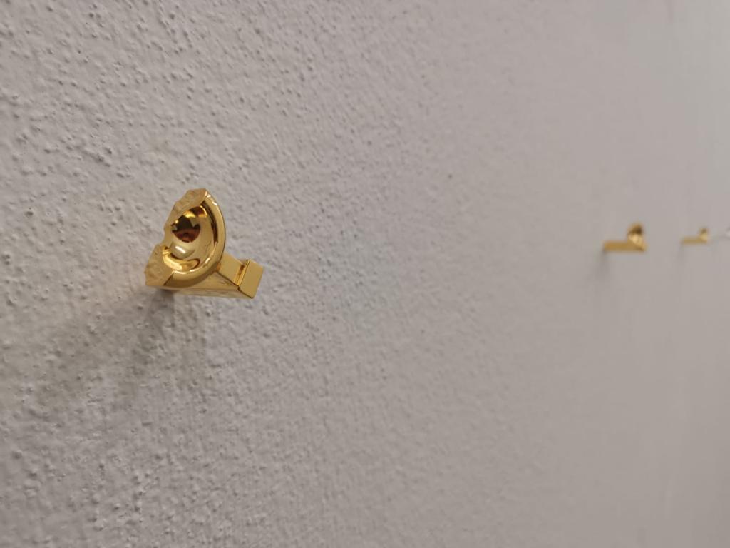 Drei kleine goldene Objekte, davon zwei nur schemenhaft zu erkennen, vor weißer Raufasertapete.