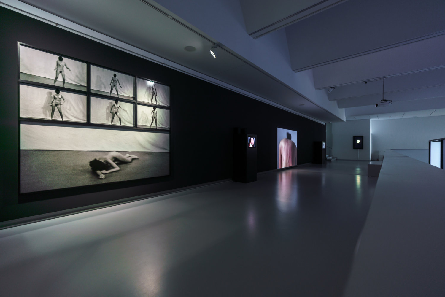 Ausstellungsansicht der Kunsthalle Tübingen, der Raum ist dunkel und es sind Performance-Videos der Künstlerin Marina Abramovic zu sehen. Im Vordergrund sieht man verschiedene Videos, wo sie nackt tanzt und zu Boden fällt, im Hintergrund sieht man ihren roten, nackten Rücken