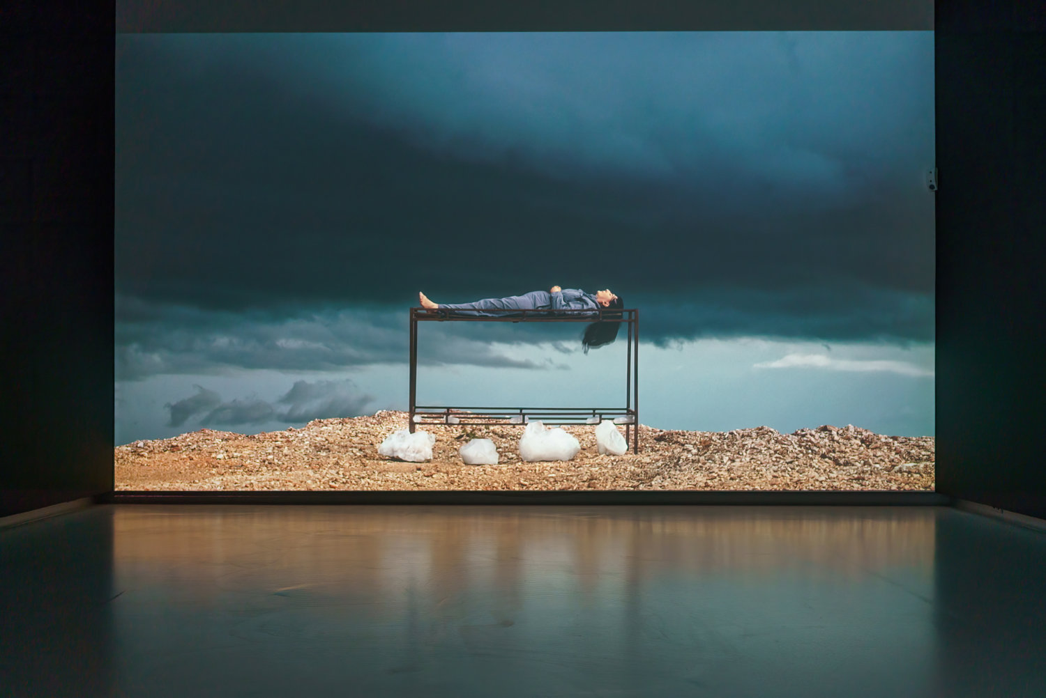 Große Videowand in einem Ausstellungsraum, die einen dunklen Himmel zeigt und eine Landschaft. Die Künstlerin Marina Abramovic liegt mit dem Rücken auf einem Gestell.