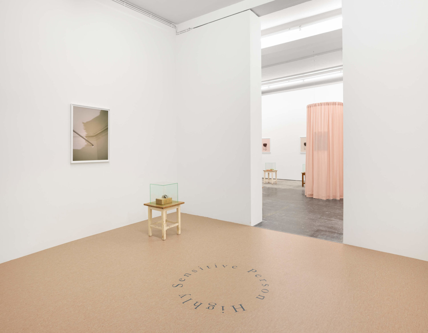 Der Blick in den Raum der Galeria Plan B zeigt einen Schriftzug auf dem Boden, einen rosa Vorhang und Fotografien an den Wänden