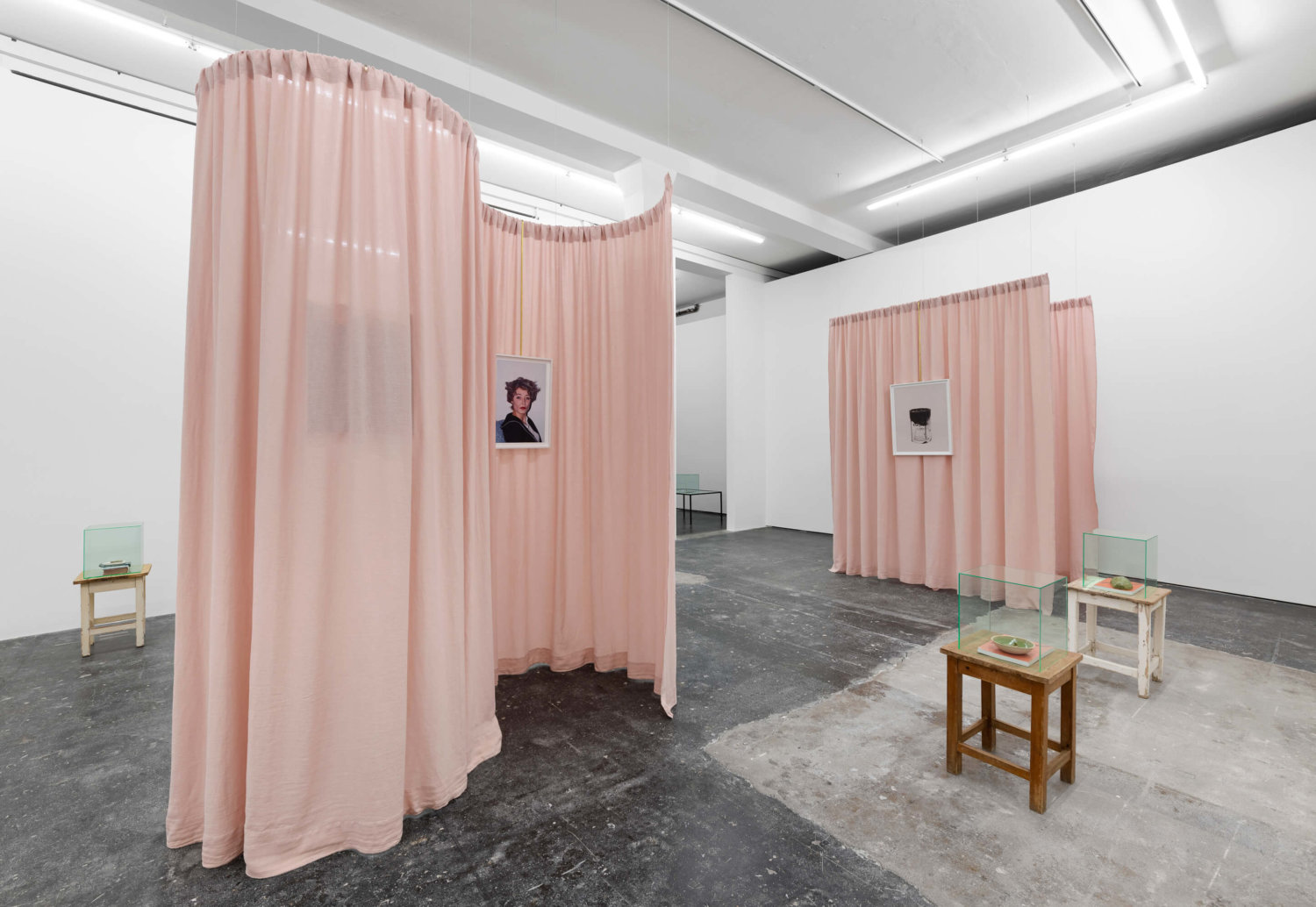 Zwei rosa Vorhänge teilen den Raum. Daran sind Fotografien befestigt. Darum herum stehen Hocker mit Objekten darauf.
