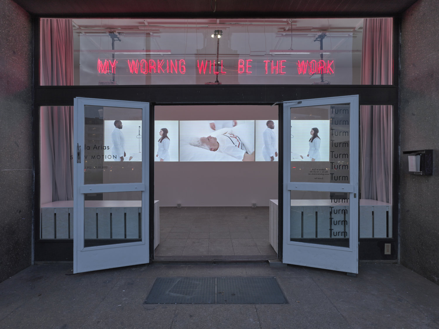 Eingang der Galerie im Turm mit Blick auf drei Videoleinwände, darüber steht in roter Neonröhren-Schrift "My Working Will Be The Work", der Titel der Ausstellungsreihe