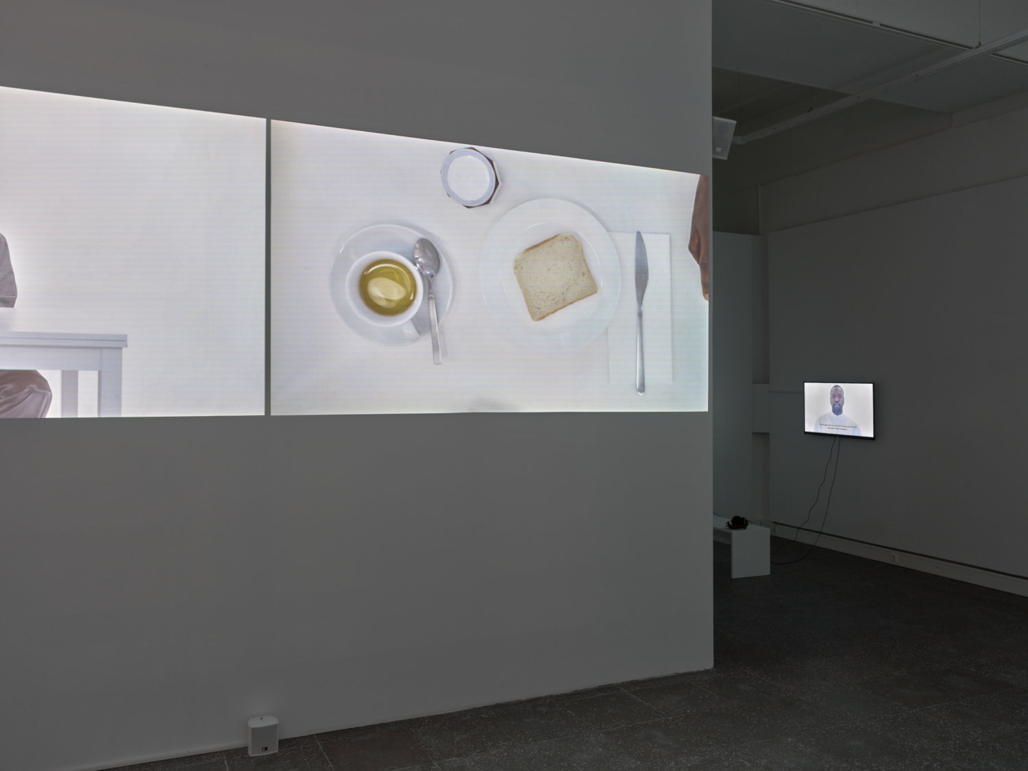 Ausstellungsansicht in der Galerie im Turm, zu sehen sind verschiedene Videoarbeiten und weiße Ausstellungswände, im Vordergrund ist ein Toast auf einem weißen Teller zu sehen, daneben ein Messer und eine Tasse Tee mit Löffel