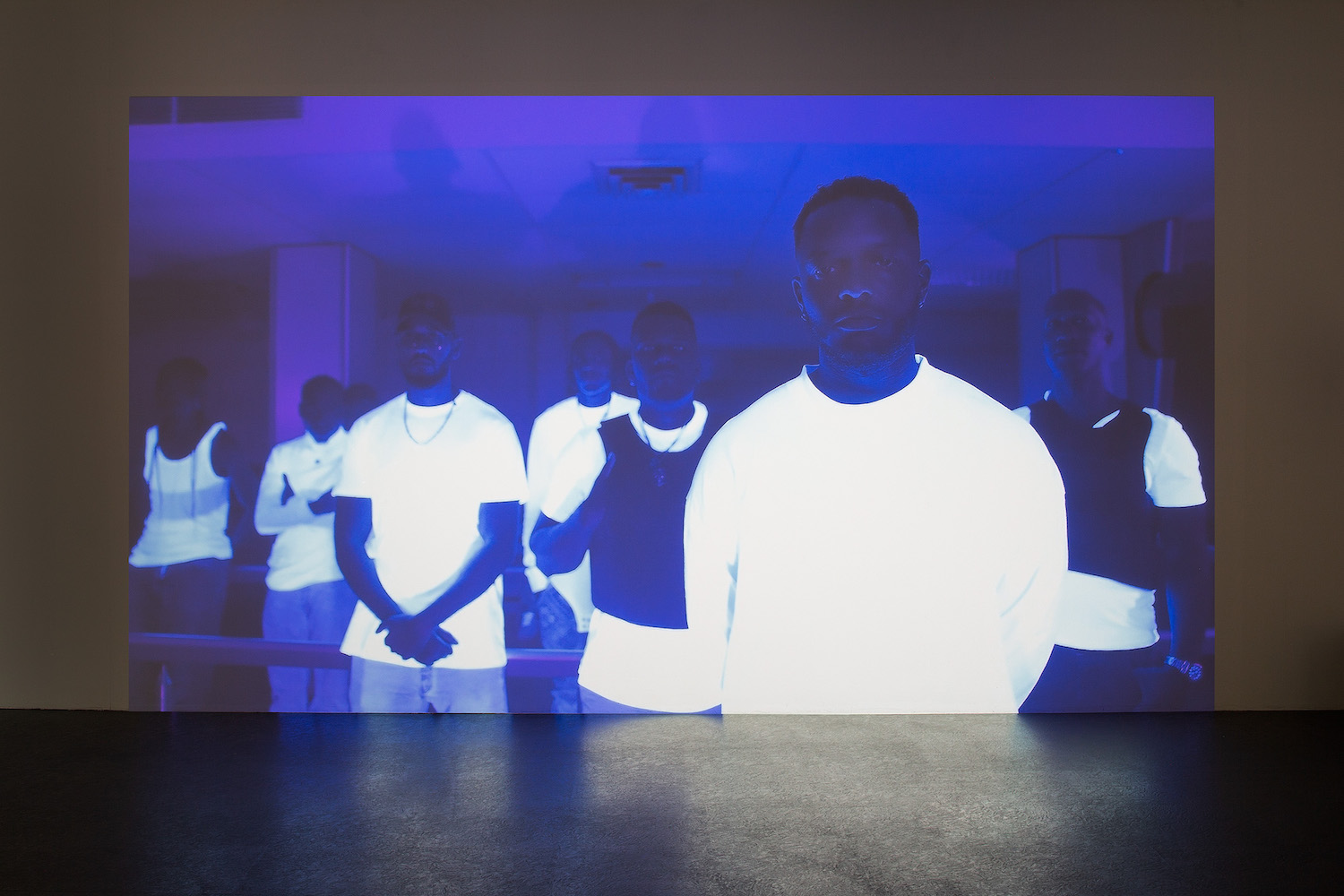 Video-Arbeit von James Gregory Atkinson. Ein Still zeigt eine Gruppe Schwarze Männer in blaues Licht getaucht. Ihre weißen Shirts leuchten, angestrahlt von Schwarzlicht.
