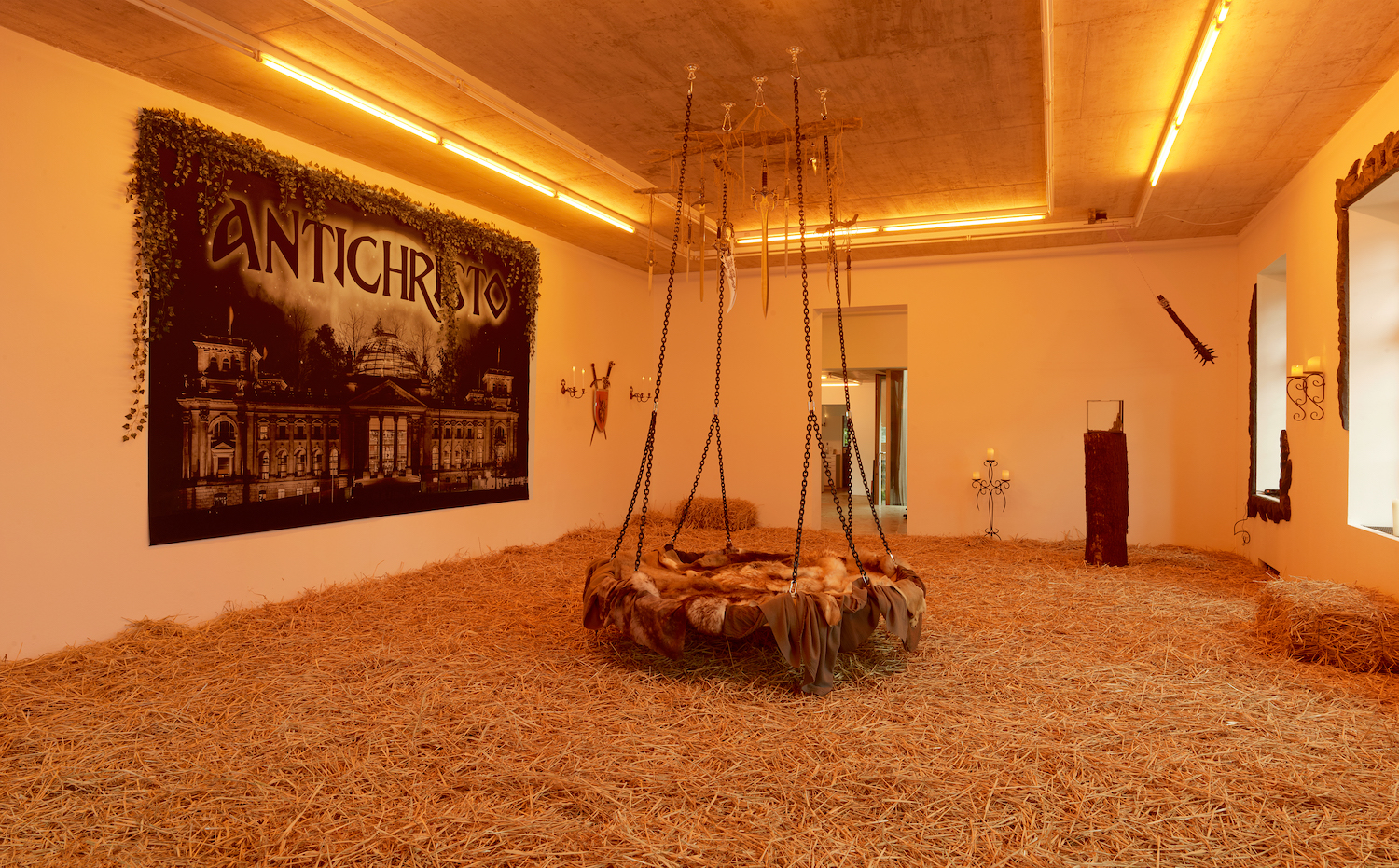 Das Erdgeschoss des Neuen Aachener Kunstvereins ist mit Stroh ausgelegt und in warmes Licht gehüllt. Zu sehen: eine fellbedeckte Schaukel mit Dolch-Mobile, ein Terrarium, um das eine Keule rotiert.