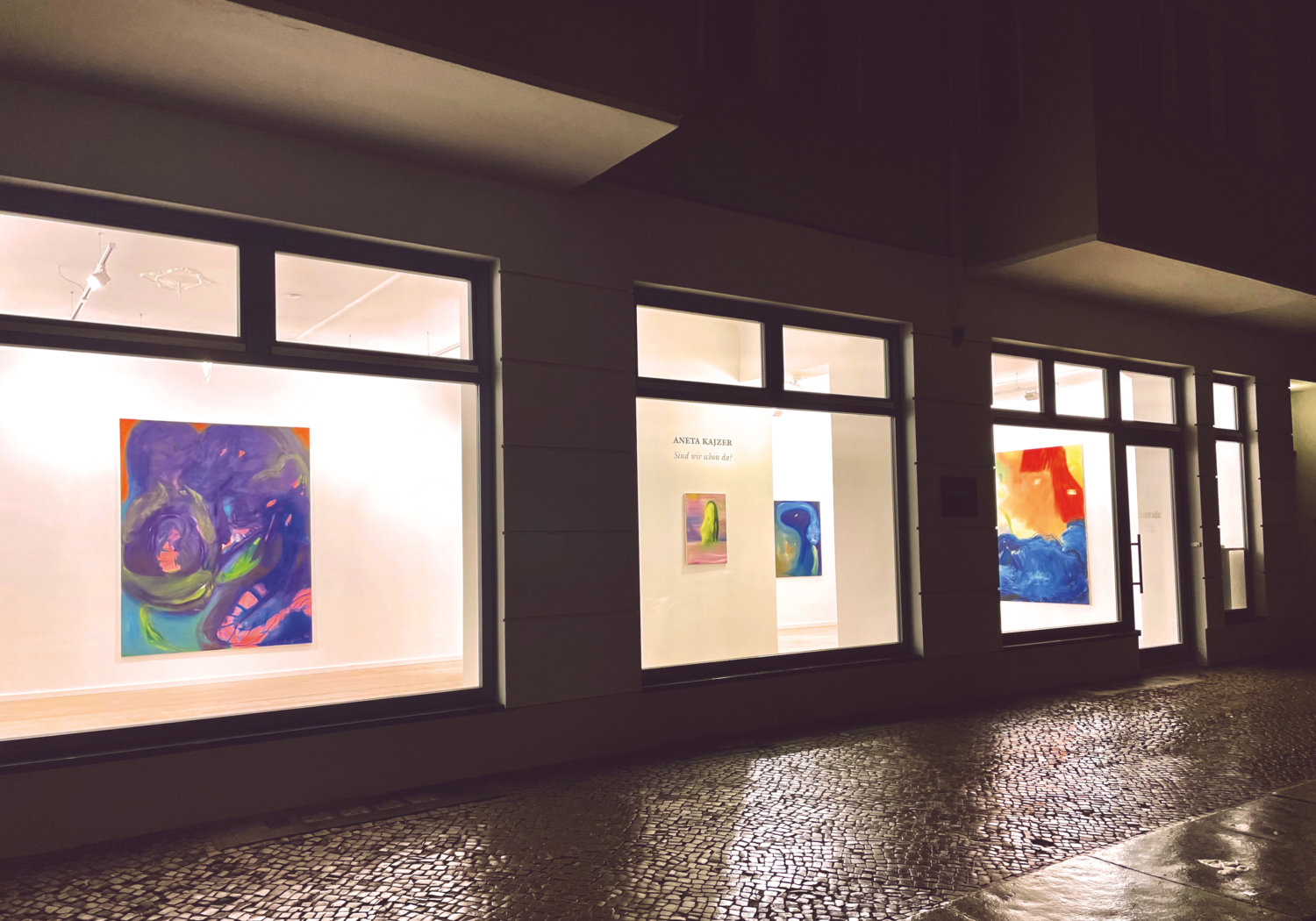 Außenansicht der Galerie Conrads mit Blick in die Ausstellung "Sind wir schon da?" von Aneta Kajzer, draußen ist es dunkel, der Ausstellungsraum hell erleuchtet