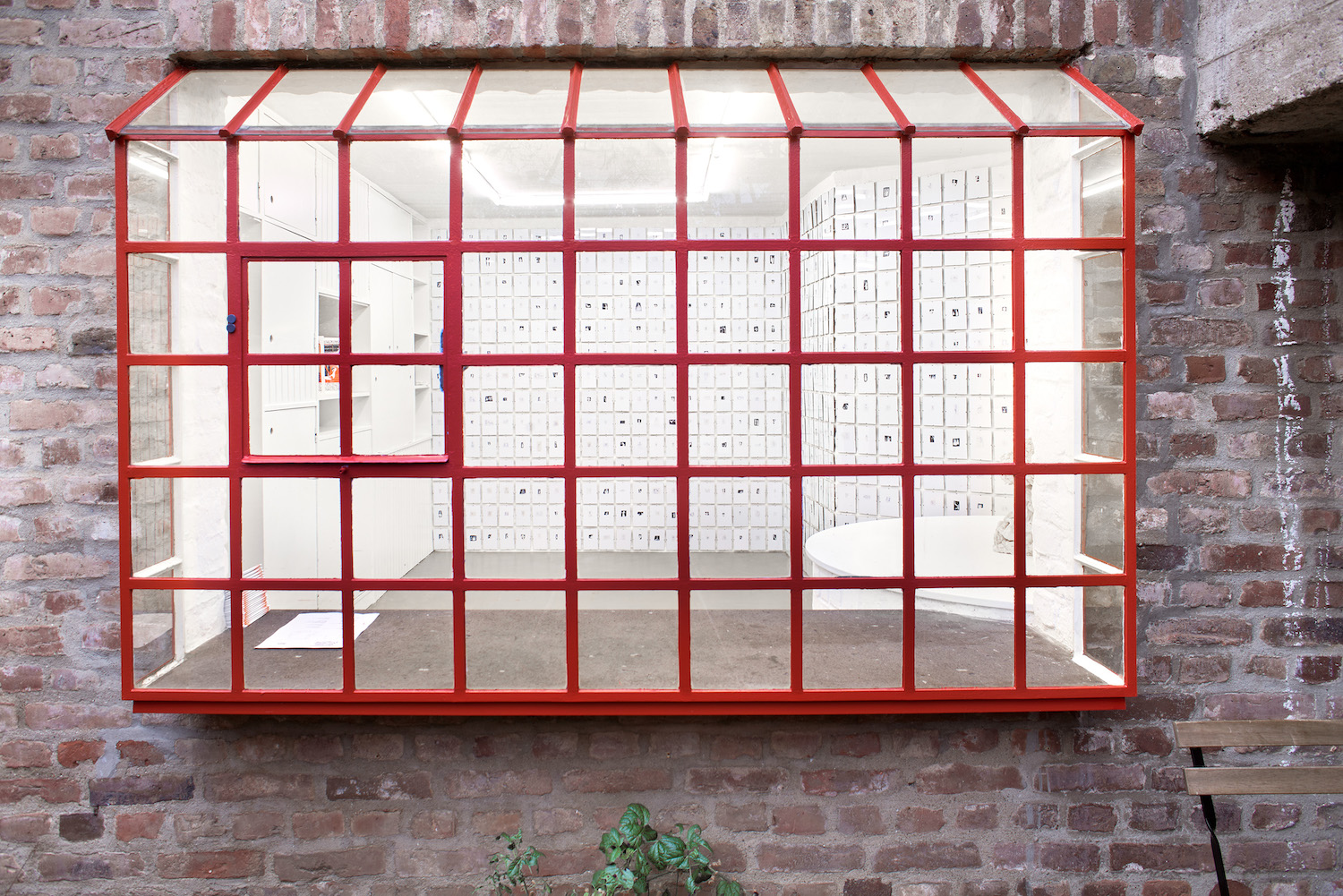 Außenansicht des Ausstellungsraums Mauer, ein großes Fenster mit roten Gitterstäben.