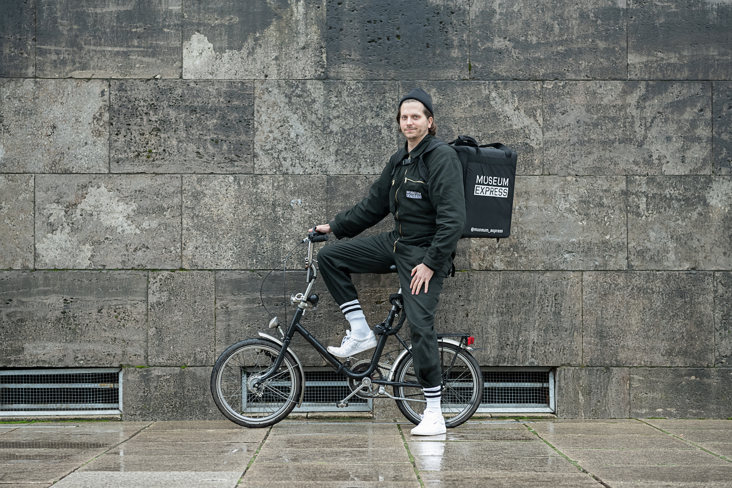 Sebastian Jung posiert auf einem Fahrrad mit dem Transportrucksack vom Museum Express auf dem Ruecken.
