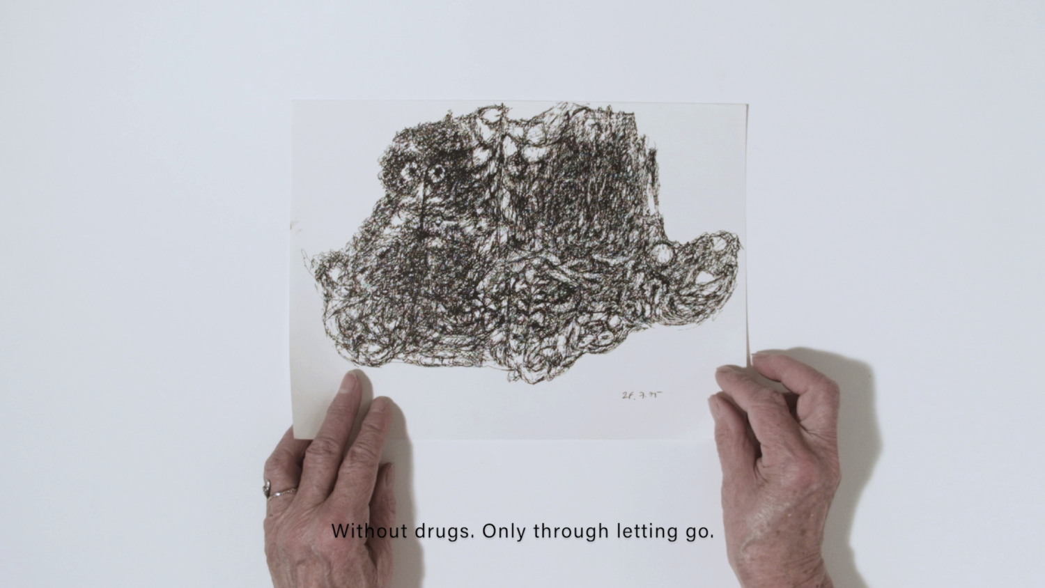 Filmstill aus "Es gibt". Man sieht Hände einer älteren Frau, die ein Papier mit einer Zeichnung halten. Darunter stehen die Worte "Without drugs. Only through letting go"
