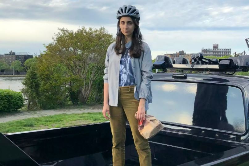 Man sieht die Künstlerin Helin Alas auf der Transportfläche eines Autos stehend, mit einem Fahrradhelm auf und einer Papiertüte in der Hand
