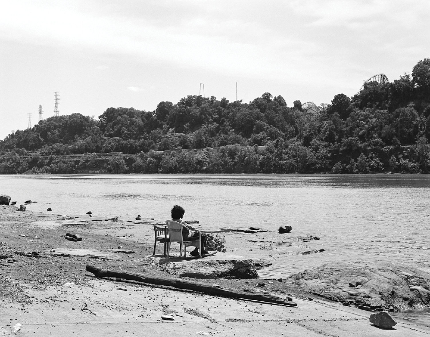 Schwarz-Weiß-Fotografie von einem Gewässer, eine Person sitzt davor auf einem Stuhl und blickt auf das Wasser, im Hintergrund sind Bäume zu sehen