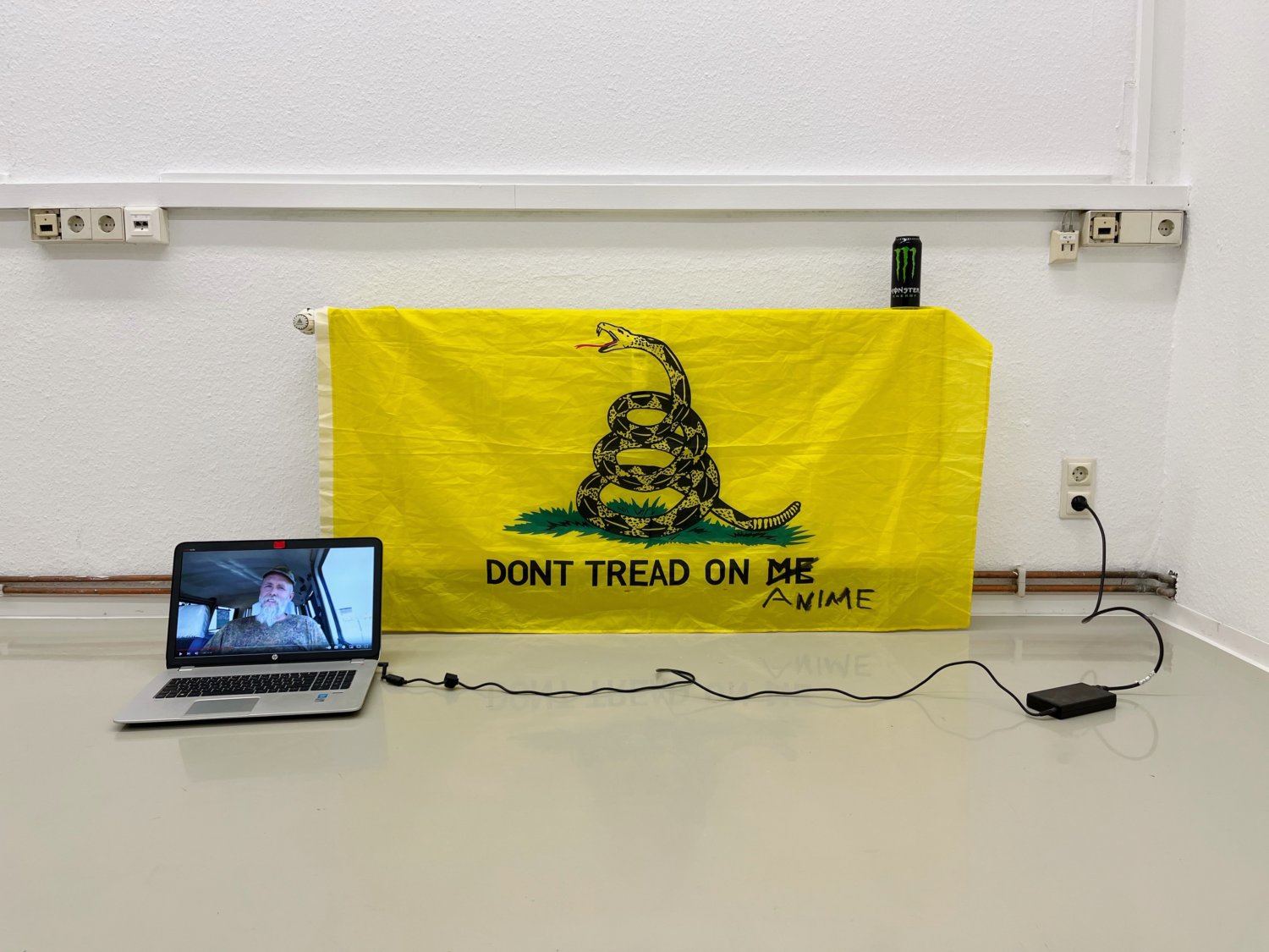 Die Arbeit "myheritage.com" von Nicolai Sauer. Eine Gadsen Flag, ein Monster Energy Drink und ein aufgeklappter Laptop, auf dem ein Video mit einem Mann in Uniform läuft.