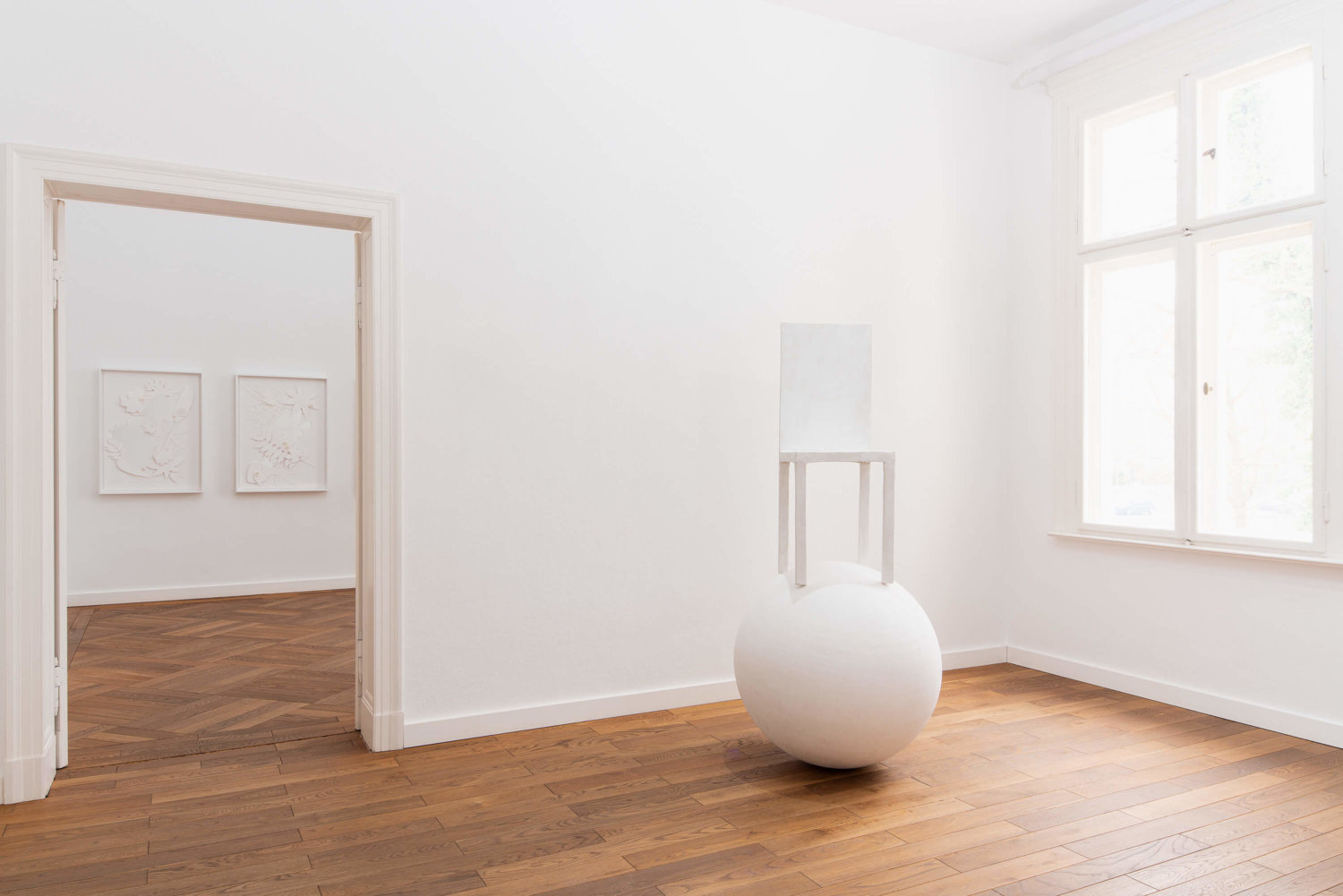 Ausstellungsansicht in der Villa Schöningen, im Vordergrund ein Stuhl, der auf einer weißen Kugeln balanciert, im Hintergrund gerahmte Papier-Arbeiten an einer weißen Wand