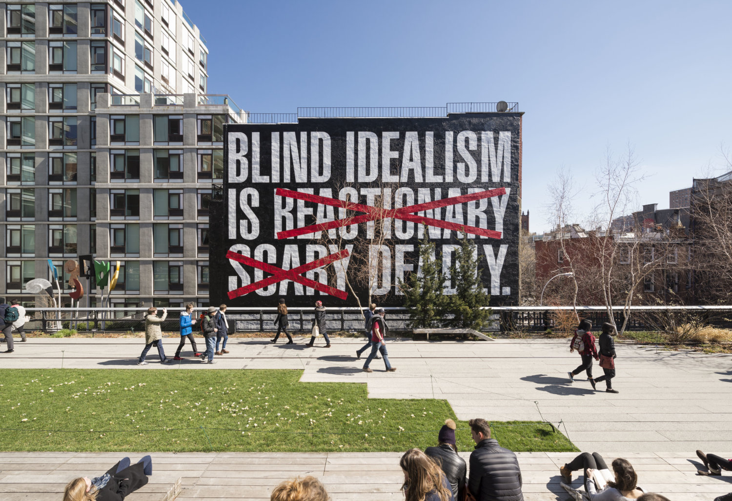 Ansicht eines öffentlichen Raums mit Wohnhäusern, einer grünen Wiese und Fußgängern, an einer Fassade ist ein großer Banner mit einem Schriftzug, zwei Worte sind in rot durchkreuzt