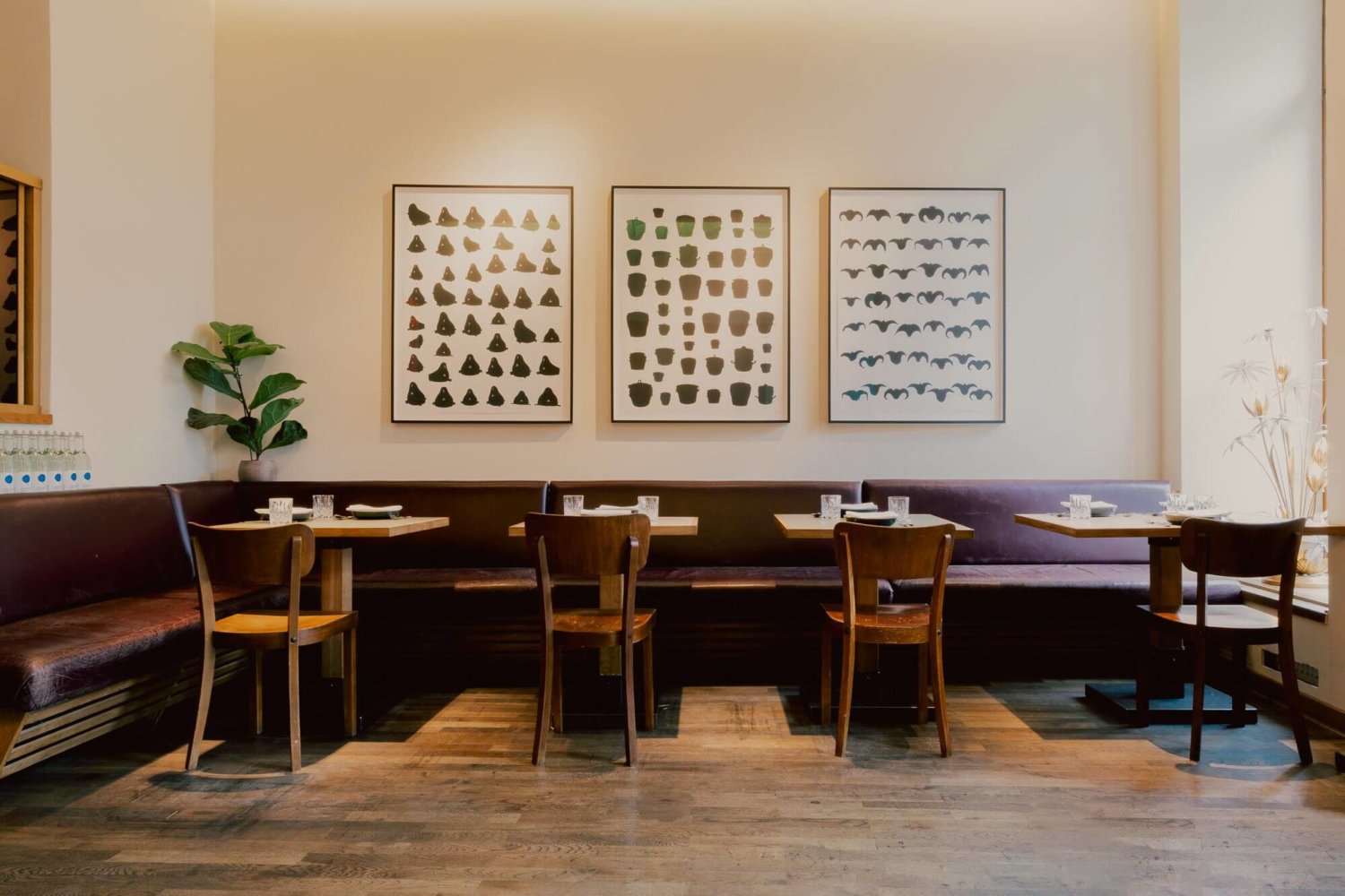 Blick in das Restaurant Kin Dee, zu sehen sind vier kleine Tische und eine lange Sitzbank sowieso einzelne Holzstühle, im Hintergrund hängen Kunstwerke an der Wand