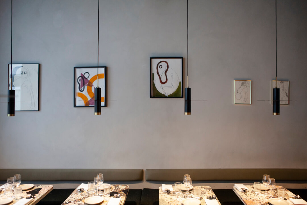 Blick an eine Wand im La Côte Restaurant, zu sehen sind kleine gedeckte Tische über denen Lampen hängen, an der Wand hängen kleinformatige Kunstwerke