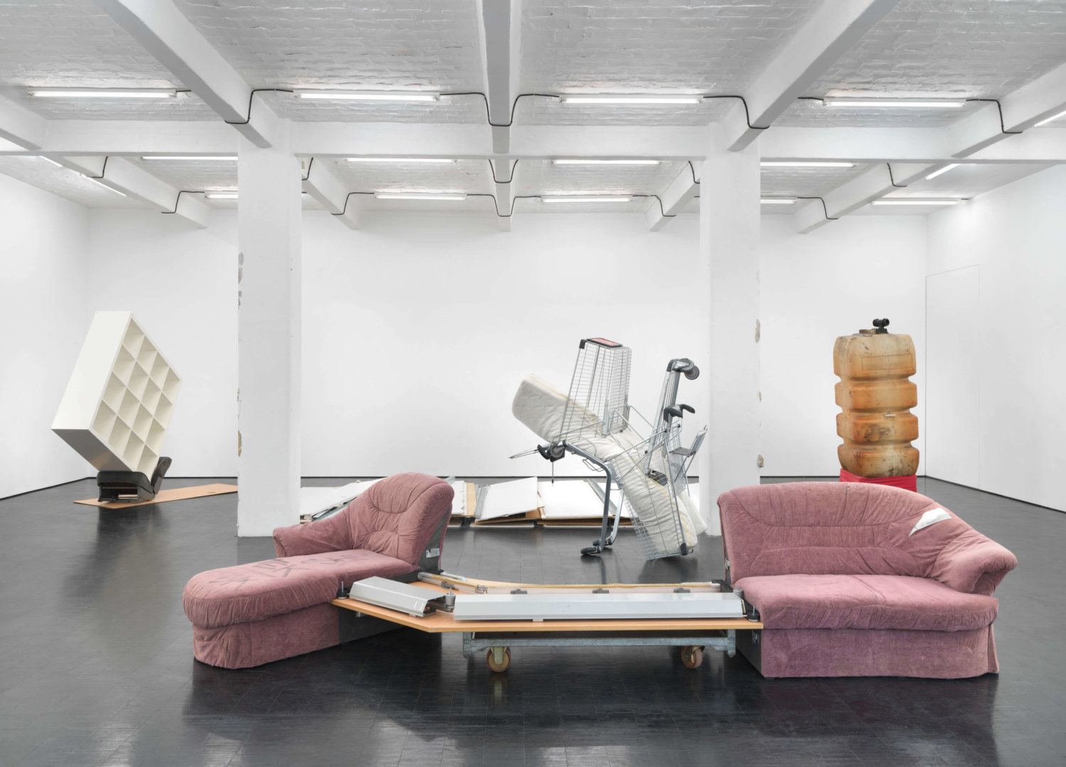 verschiedene Objekte im Ausstellungsraum: ein zweigeteiltes Sofa, Einkaufwägen, ein Regal, ein großer Kanister