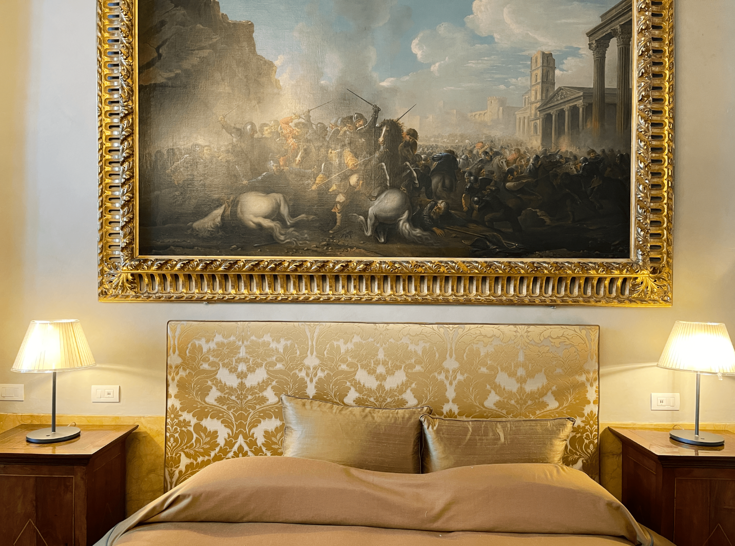 Ansicht eines Hotelzimmers und einem Bett, daneben zwei Nachtische mit Lampen und über dem Bild hängt ein großes Gemälde, fast alles im Bildausschnitt ist goldfarben