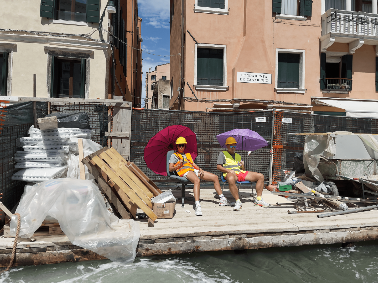 Frau und Mann sitzen in Neapel auf einer Baustelle direkt am Wasser aus zwei Stühlen, tragen Warnwesten und Schutzhelme und bunte Regenschirme als Sonnenschutz