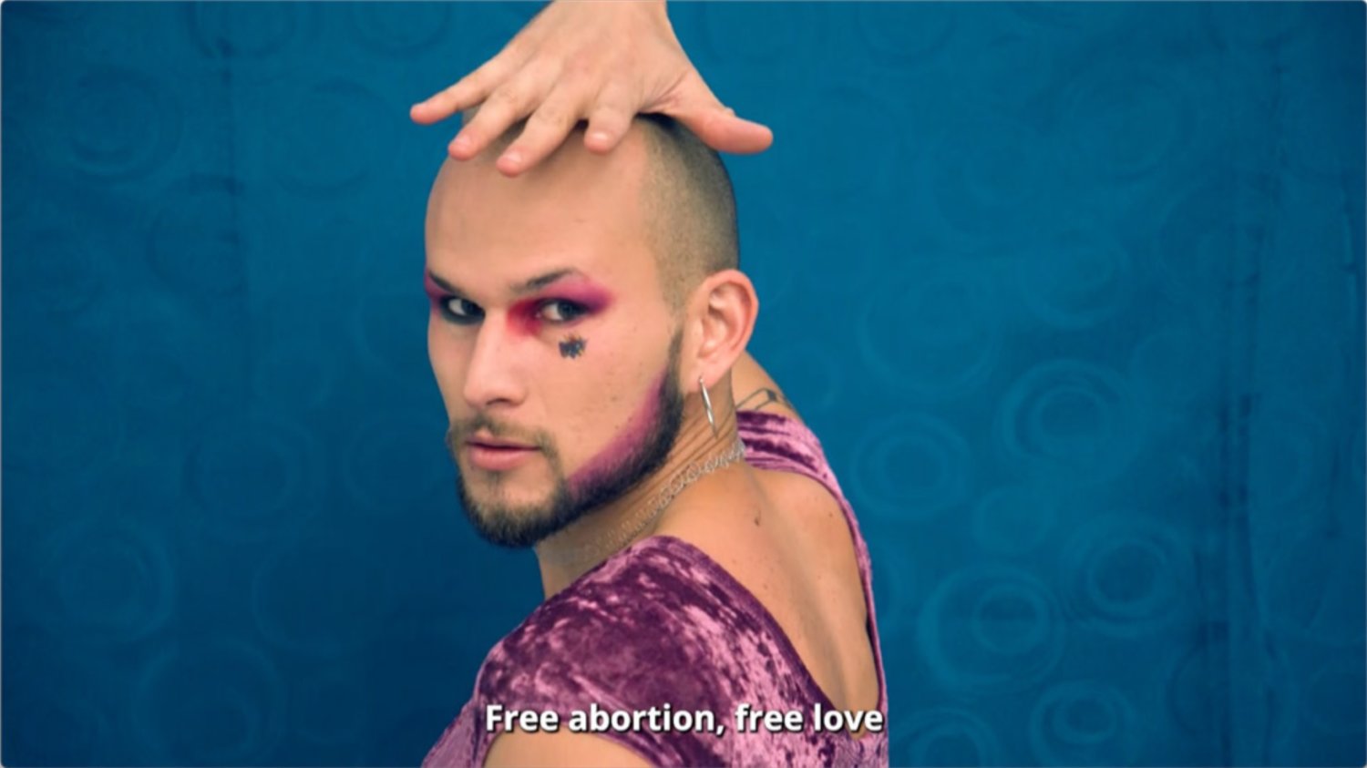 Eine geschminkte männlich gelesene Person im Samtoberteil guckt in die Kamera. Darunter steht "Free abortion, free love".