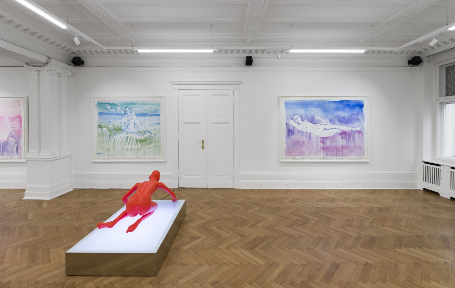 An der Wand hängen zwei große Aquarellmalereien von Marianna Simnett. Auf dem Boden steht eine Skulptur der Künstlerin, die komplett rot ist und einen nackten Körper zeigt.